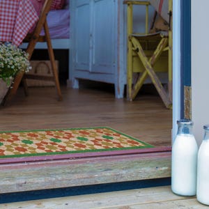 May include: A rectangular doormat with a geometric pattern of orange, green, and tan diamonds on a natural fiber background. The mat is framed with a green border. Two glass milk bottles are in the foreground. The mat is on a wooden porch.