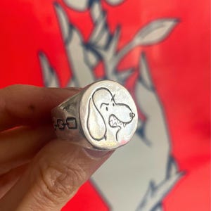 May include: A silver ring with a round, engraved design of a cartoon dog with a surprised expression. The dog's mouth is open, showing teeth.