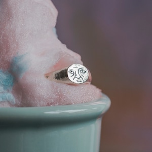 May include: A silver ring with a face design on the top. The ring is sitting in a light blue bowl filled with pink candy floss.