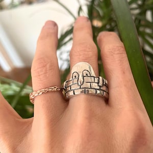 May include: A silver ring with a design of a face peeking over a brick wall. The ring is worn on a person's finger.