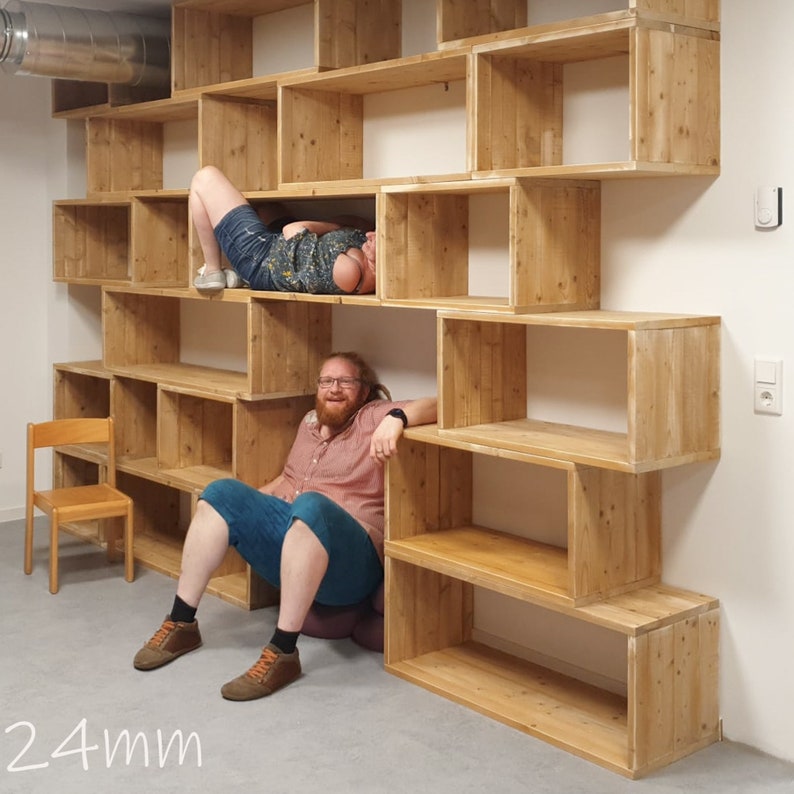 Puede incluir: Una estanter&iacute;a de madera con varios estantes de diferentes tama&ntilde;os. Los estantes est&aacute;n hechos de madera natural y tienen un aspecto r&uacute;stico. Dos personas est&aacute;n sentadas en los estantes, una est&aacute; tumbada y la otra est&aacute; sentada. Los estantes est&aacute;n contra una pared blanca.