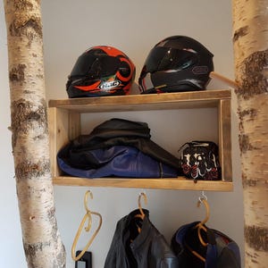 May include: A rectangular wooden wall shelf mounted between two birch tree trunks. The shelf holds two motorcycle helmets, a folded blue and black jacket, and a pair of gloves. Below the shelf, three hangers hold motorcycle jackets.