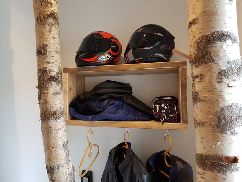 May include: A rectangular wooden wall shelf mounted between two birch tree trunks. The shelf holds two motorcycle helmets, a folded blue and black jacket, and a pair of gloves. Below the shelf, three hangers hold motorcycle jackets.