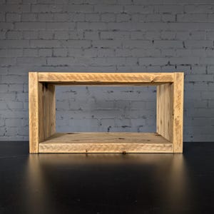 May include: A rectangular wooden shelf unit constructed from light brown reclaimed timber with a visible grain pattern. The structure features two vertical side panels supporting a horizontal top and bottom board, creating an open storage space.