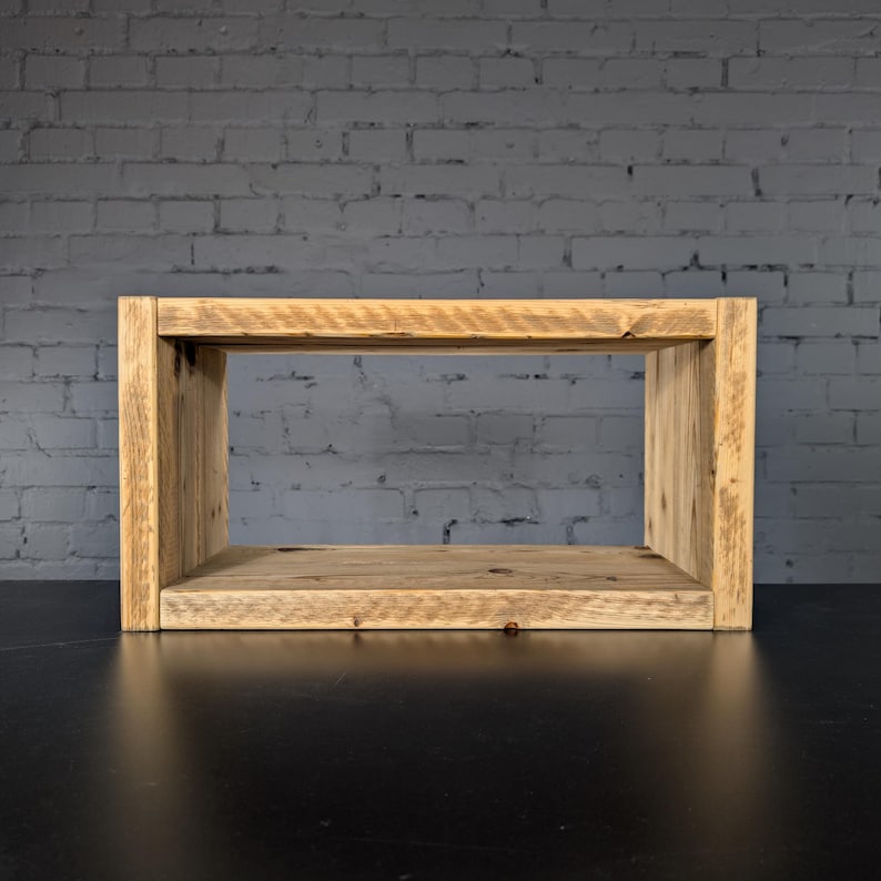 May include: A rectangular wooden shelf unit constructed from light brown reclaimed timber with a visible grain pattern. The structure features two vertical side panels supporting a horizontal top and bottom board, creating an open storage space.