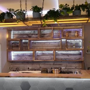 May include: A wall-mounted wooden shelving unit with multiple rectangular compartments and integrated LED strip lighting. The shelves are constructed from weathered planks and feature various decorative items, including metallic pitchers and a glass bottle.