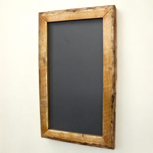 Puede incluir: Pizarra rectangular con una superficie gris oscuro, enmarcada por un borde de madera marr&oacute;n claro r&uacute;stico. La madera tiene vetas y nudos visibles, lo que le da un aspecto natural y texturizado. La pizarra est&aacute; montada en una pared blanca.