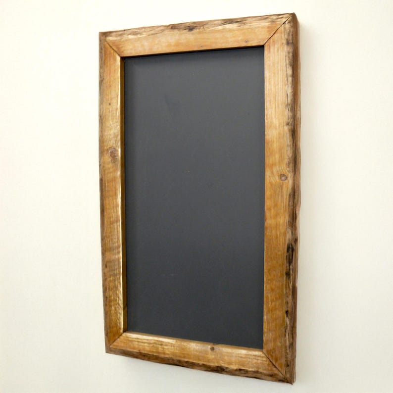 Puede incluir: Pizarra rectangular con una superficie gris oscuro, enmarcada por un borde de madera marr&oacute;n claro r&uacute;stico. La madera tiene vetas y nudos visibles, lo que le da un aspecto natural y texturizado. La pizarra est&aacute; montada en una pared blanca.