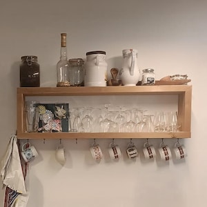 May include: A two-tiered wooden wall shelf featuring a top surface holding glass jars, a ceramic pitcher, and a wooden scoop. The lower shelf contains various clear drinking glasses, while metal hooks underneath display several patterned mugs.