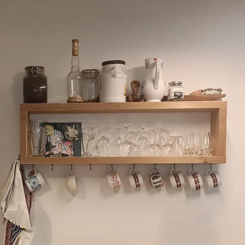 May include: A two-tiered wooden wall shelf featuring a top surface holding glass jars, a ceramic pitcher, and a wooden scoop. The lower shelf contains various clear drinking glasses, while metal hooks underneath display several patterned mugs.