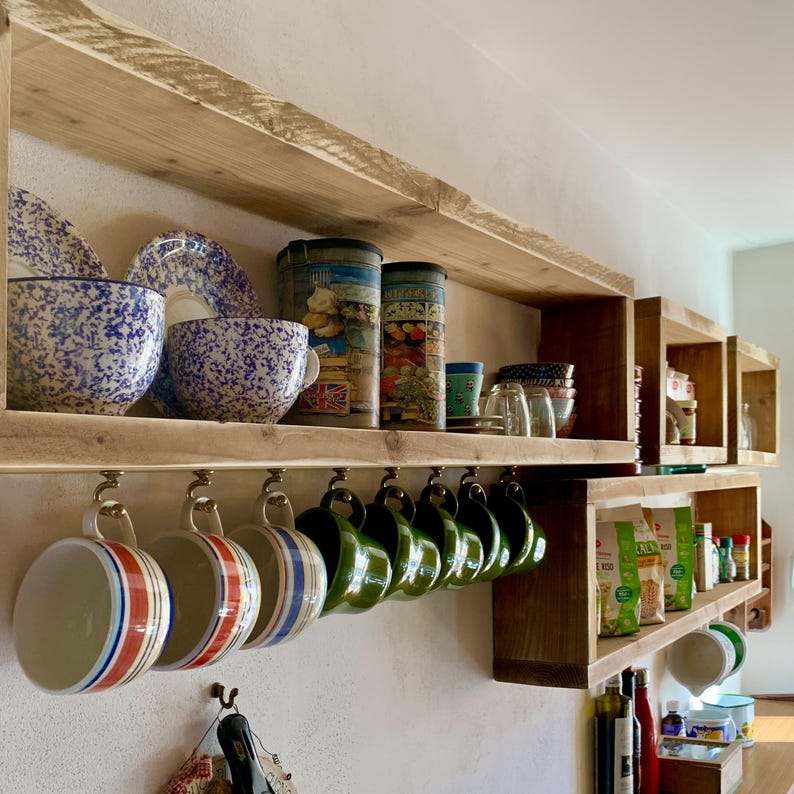 May include: Natural wood floating shelves mounted on a wall, featuring blue and white speckled bowls, decorative canisters, and a row of hanging ceramic mugs with red, blue, and green striped patterns, plus additional kitchen storage items.
