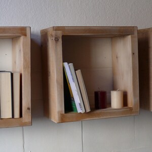May include: Three wooden wall shelves with a rustic finish. The shelves are empty except for the middle shelf, which has a stack of books and two candles.