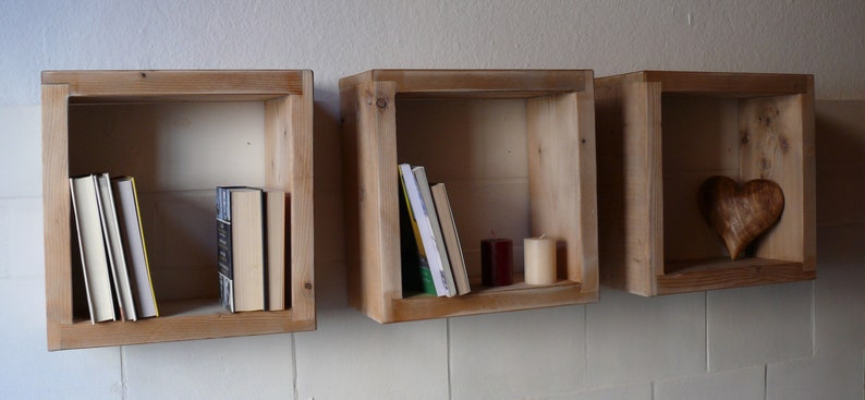 May include: Three wooden wall shelves with a rustic finish. The shelves are empty except for the middle shelf, which has a stack of books and two candles.