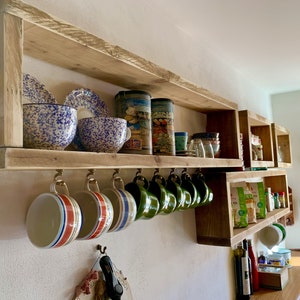 Könnte beinhalten: Ein wandmontiertes Holzregal mit natürlichem Finish, das auf der oberen Ablage blau-weiß gesprenkelte Schalen und Vorratsdosen sowie an einer unteren Reihe von Messinghaken eine Sammlung bunter gestreifter und grüner Tassen präsentiert.