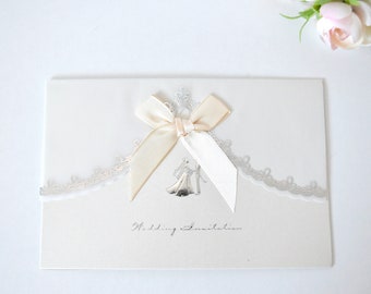 May include: A white wedding invitation with a silver lace border and a cream satin bow. The invitation says "Wedding Invitation" in cursive.