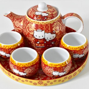 May include: A red and yellow Hello Kitty tea set. The set includes a teapot, four cups, and a tray. The teapot and cups feature Hello Kitty designs and floral patterns. The tray has a floral border. The set is designed for serving tea.