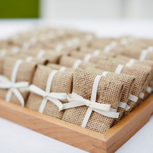 May include: A wooden tray holding numerous small, rectangular packages wrapped in burlap and tied with white ribbon bows. The packages are arranged in rows, possibly as wedding favours or small gifts, with a light background.