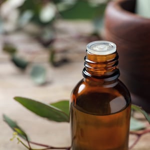 Puede incluir: Una botella de vidrio marrón con una tapa blanca llena de un líquido transparente. La botella está sobre una superficie de madera con hojas verdes.