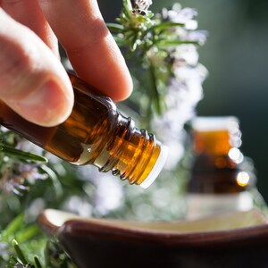 May include: A brown glass essential oil bottle is being poured into a small brown ceramic bowl. The bottle has a white cap and is surrounded by green and purple plants. The background is blurred, with hints of green.