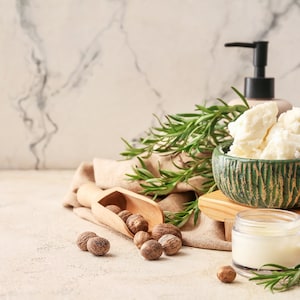 May include: A green bowl filled with white shea butter, a small jar of cream, a wooden scoop filled with nutmeg, and sprigs of rosemary on a light brown surface.