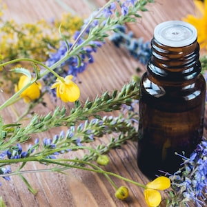 May include: A brown glass bottle with a white lid filled with essential oil, surrounded by purple and yellow wildflowers on a wooden surface.