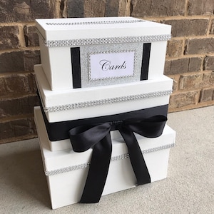 May include: A three-tiered white gift box with black ribbon and silver glitter trim. The top box has a silver glitter label that says "Cards".