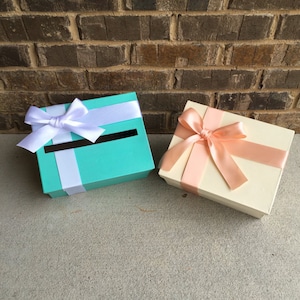 May include: Two gift boxes with lids, one is teal with a white ribbon and bow, the other is cream with a peach ribbon and bow.