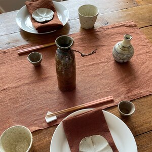 May include: A table setting with two place settings, each with a white plate, brown linen napkin, and a small white ceramic dish. There are two small brown ceramic cups, a brown ceramic bottle, and a small white ceramic vase with a brown and white pattern.
