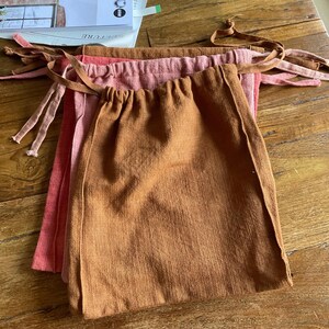 Op de afbeelding: Drie stoffen trekzakken in verschillende tinten bruin, roze en rood. De zakken zijn gemaakt van een zacht, natuurlijk materiaal en hebben een eenvoudig, rustiek ontwerp.