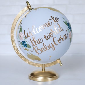 Puede incluir: Un globo terr&aacute;queo azul con un soporte dorado. El globo est&aacute; decorado con pintura dorada, flores y las palabras "Bienvenido al mundo, &iexcl;Beb&eacute; Corsi!"