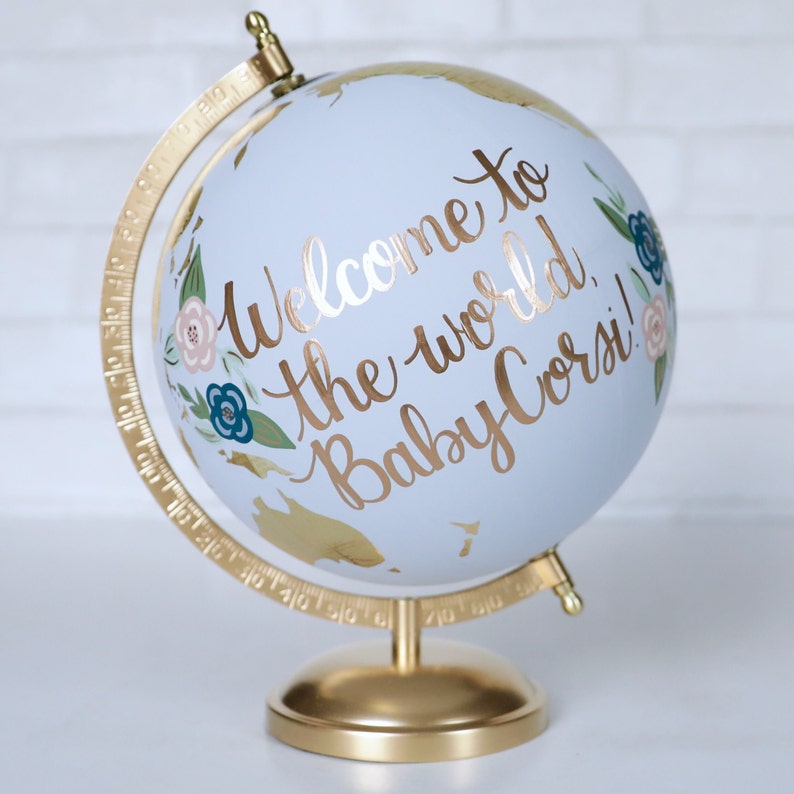 Puede incluir: Un globo terr&aacute;queo azul con un soporte dorado. El globo est&aacute; decorado con pintura dorada, flores y las palabras "Bienvenido al mundo, &iexcl;Beb&eacute; Corsi!"