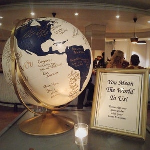 Peut inclure: Un globe terrestre blanc et bleu avec des accents dorés est utilisé comme livre d'or lors d'une réception de mariage. Le globe est décoré de messages et de signatures manuscrits. Une pancarte à côté du globe indique "You Mean The World To Us! Please sign our guest globe with your name & wishes."