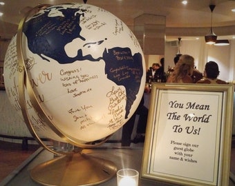 Puede incluir: Un globo terráqueo blanco y azul con detalles dorados se utiliza como libro de visitas en una recepción de boda. El globo está decorado con mensajes y firmas escritas a mano. Un cartel junto al globo dice "You Mean The World To Us! Please sign our guest globe with your name & wishes."