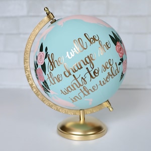 Puede incluir: Un globo de color azul claro con un soporte dorado. El globo est&aacute; pintado con detalles florales de color rosa y verde y las palabras "She will be the change she wants to see in the world".