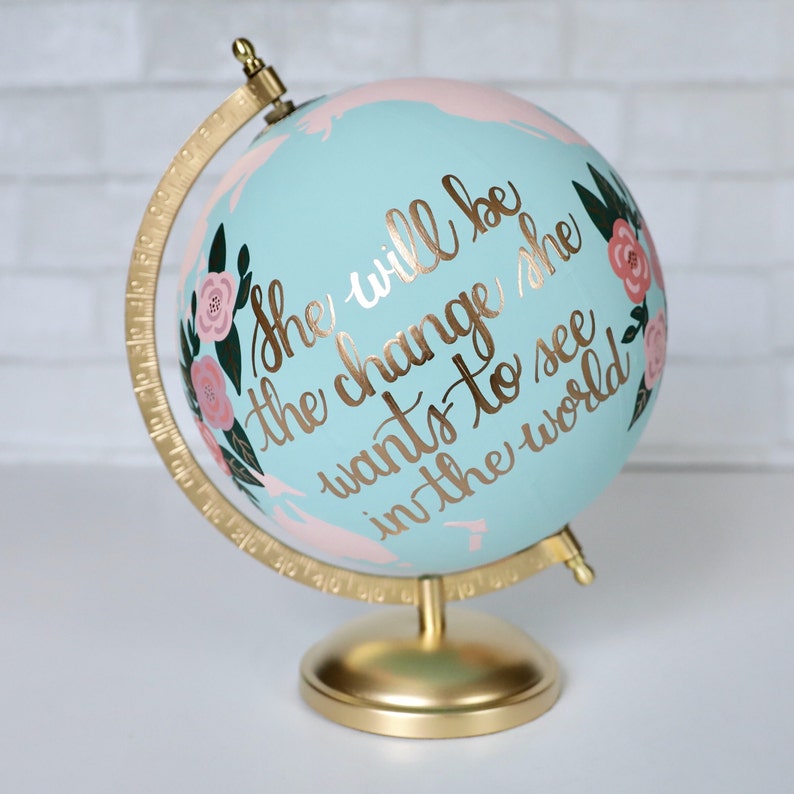 Puede incluir: Un globo de color azul claro con un soporte dorado. El globo est&aacute; pintado con detalles florales de color rosa y verde y las palabras "She will be the change she wants to see in the world".