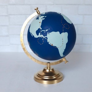 Puede incluir: Un globo terr&aacute;queo azul con un soporte dorado. El globo tiene un mapa del mundo azul claro con l&iacute;neas doradas y corazones.