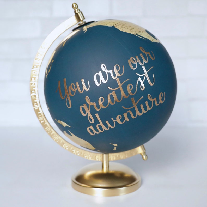 Puede incluir: Un globo terr&aacute;queo azul con un soporte dorado. El globo tiene una inscripci&oacute;n dorada que dice "You are our greatest adventure".