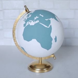 Puede incluir: Un globo terr&aacute;queo blanco con un mapa del mundo azul turquesa. El globo tiene un soporte dorado y un meridiano dorado.