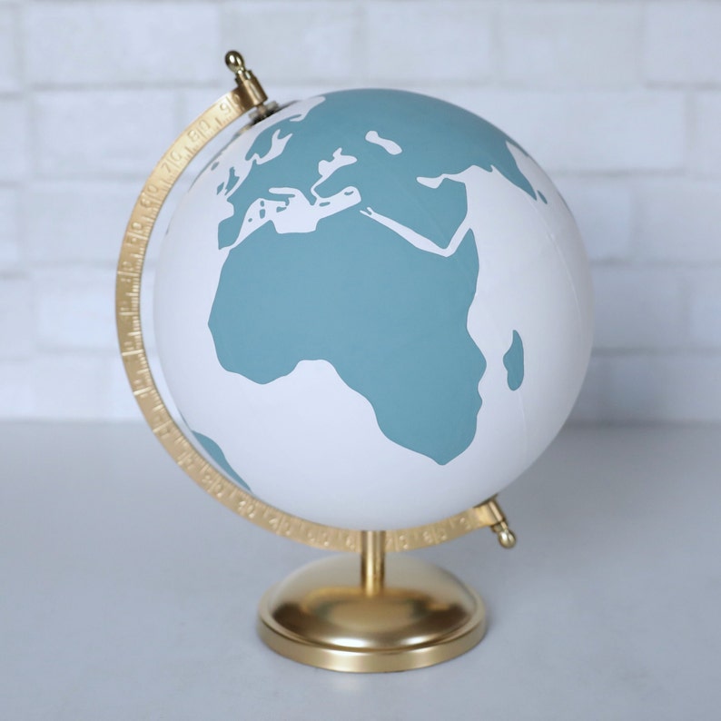 Puede incluir: Un globo terr&aacute;queo blanco con un mapa del mundo azul turquesa. El globo tiene un soporte dorado y un meridiano dorado.