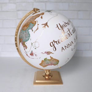 Peut inclure: Globe blanc avec un socle doré. Le globe a une carte du monde imprimée dessus et un avion doré avec une ligne pointillée. Le globe est personnalisé avec le texte "You're my greatest adventure" et "Anna & Brian 6.21.19".