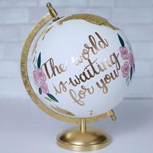 Puede incluir: Un globo terr&aacute;queo blanco con una base y un meridiano dorados. El globo est&aacute; decorado con flores rosas y hojas verdes. El texto "The world is waiting for you" est&aacute; escrito en letras doradas en el globo.