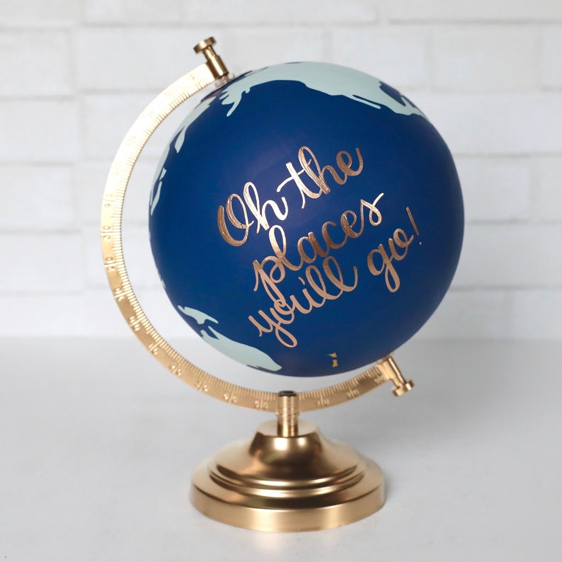 Puede incluir: Un globo terr&aacute;queo azul con un soporte dorado. El globo tiene un mapa del mundo blanco y las palabras "Oh the places you'll go!" escritas en letras doradas.