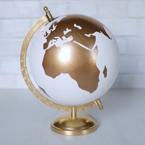 Puede incluir: Un globo terr&aacute;queo dorado y blanco con un soporte dorado. El globo tiene un fondo blanco con continentes dorados.
