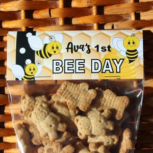 May include: A clear plastic bag filled with small, brown, bear-shaped cookies. The bag has a yellow and black label with the text "Ava's 1st BEE DAY" and a cartoon bee.