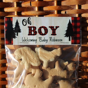May include: A clear plastic bag filled with animal crackers. The bag has a label that says "Oh Boy" in red and black letters with a red and black buffalo plaid background. The label also says "Welcoming Baby Robinson".