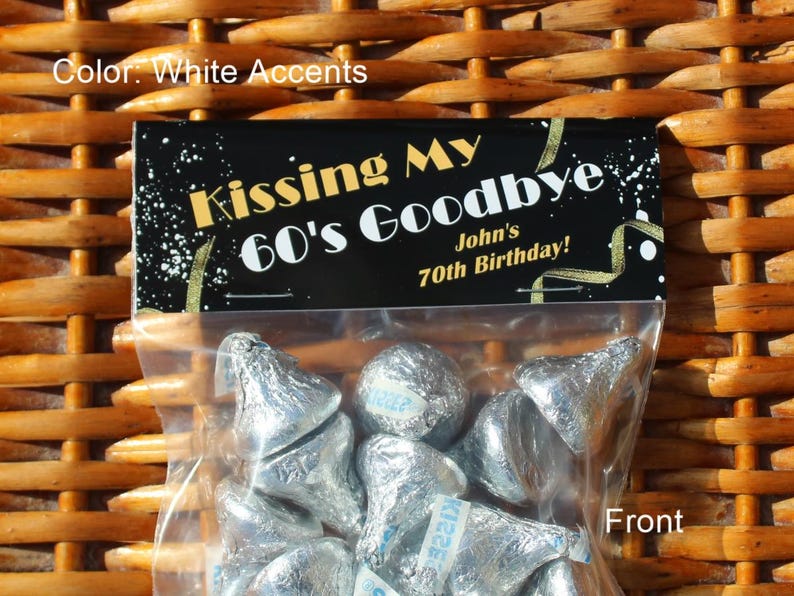 May include: A black and gold party favour bag with the text "Kissing My 60's Goodbye John's 70th Birthday!" The bag is filled with silver foil-wrapped chocolate kisses.