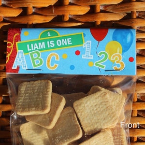 May include: A clear plastic bag filled with shortbread cookies. The bag has a colorful label that says "Liam is One" with the letters ABC and the numbers 123. The label also has red, yellow, and blue balloons.