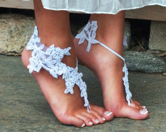 May include: White lace barefoot sandals with floral designs. The sandals wrap around the ankles and toes, creating a delicate, feminine look. The sandals are worn on a stone surface.