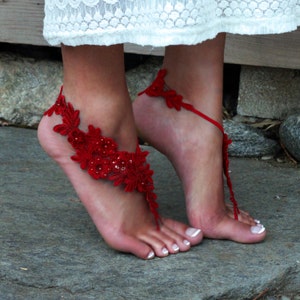 May include: Red lace barefoot sandals with beaded embellishments. The sandals are tied around the ankle and have a delicate floral design.