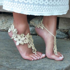 May include: A pair of ivory lace barefoot sandals with floral designs. The sandals are tied around the ankle with a ribbon.